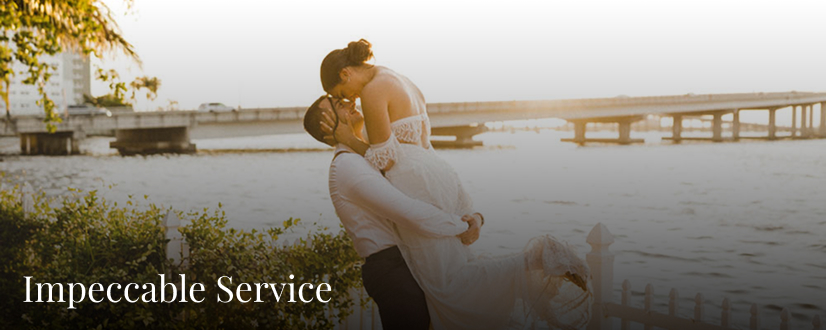 A groom lifts and kisses his bride near a waterfront at sunset, with a bridge in the background at Heitman House, an elegant waterfront wedding venue. The words Impeccable Service appear in the lower left corner. A groom lifts and kisses his bride near a waterfront at sunset, with a bridge in the background at Heitman House, an elegant waterfront wedding venue. The words Impeccable Service appear in the lower left corner.
