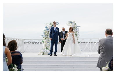 A bride and groom stand holding hands with an officiant under a floral arch on an outdoor platform by the water at Heitman House, a stunning waterfront wedding venue, while guests watch and take photos. A bride and groom stand holding hands with an officiant under a floral arch on an outdoor platform by the water at Heitman House, a stunning waterfront wedding venue, while guests watch and take photos.