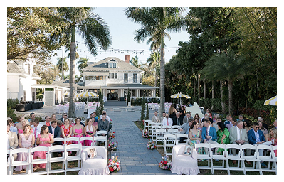 Outdoor wedding ceremony setup at Heitman House, a waterfront wedding venue with white chairs, flower-lined aisle, tall palm trees, and a large white house in the background under clear skies. Outdoor wedding ceremony setup at Heitman House, a waterfront wedding venue with white chairs, flower-lined aisle, tall palm trees, and a large white house in the background under clear skies.