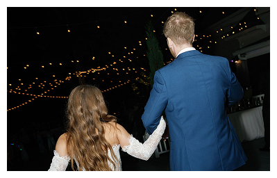 A bride and groom, seen from behind, walk together under string lights at an outdoor nighttime event at a stunning waterfront wedding venue. The bride wears a white dress and the groom wears a blue suit. A bride and groom, seen from behind, walk together under string lights at an outdoor nighttime event at a stunning waterfront wedding venue. The bride wears a white dress and the groom wears a blue suit.