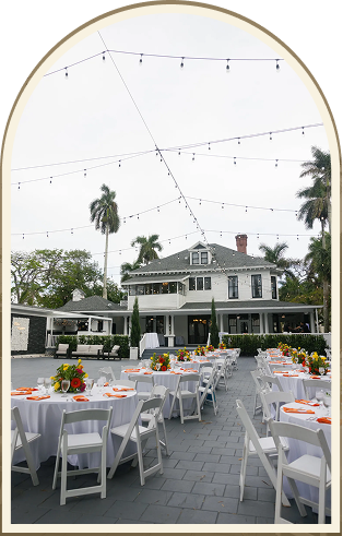 Outdoor event setup at the Heitman House, the best Fort Myers wedding venue, featuring round tables with white cloths, white chairs, colorful floral centerpieces, string lights overhead, and palm trees in the background. Outdoor event setup at the Heitman House, the best Fort Myers wedding venue, featuring round tables with white cloths, white chairs, colorful floral centerpieces, string lights overhead, and palm trees in the background.