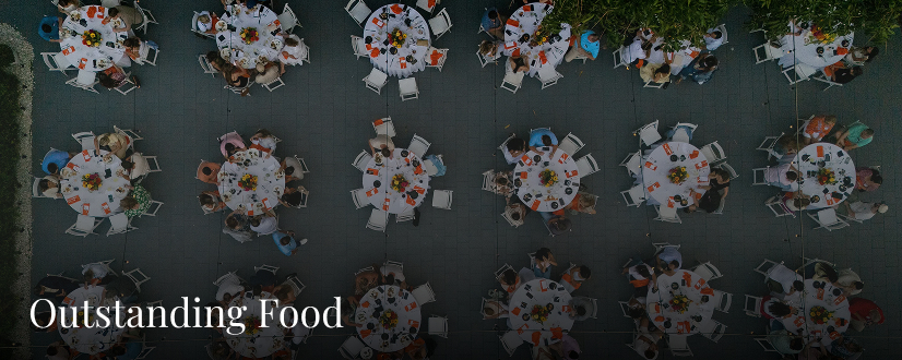 Aerial view of round tables with white tablecloths and floral centerpieces at Heitman House, one of the best Fort Myers wedding venues, surrounded by people dining outdoors. The text Outstanding Food appears at the bottom left. Aerial view of round tables with white tablecloths and floral centerpieces at Heitman House, one of the best Fort Myers wedding venues, surrounded by people dining outdoors. The text Outstanding Food appears at the bottom left.