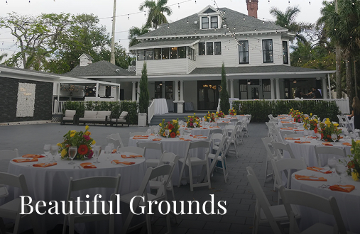 Outdoor event setup with round tables and white chairs, decorated with colorful flower centerpieces, in front of the elegant Heitman House. String lights hang above this stunning waterfront wedding venue. Beautiful Grounds is written at the bottom. Outdoor event setup with round tables and white chairs, decorated with colorful flower centerpieces, in front of the elegant Heitman House. String lights hang above this stunning waterfront wedding venue. Beautiful Grounds is written at the bottom.
