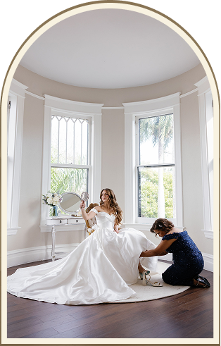 A bride in a white gown sits on a chair in a bright, round room at Heitman House, the best Fort Myers wedding venue, as another woman kneels to help adjust her dress. Large windows and a vanity with a mirror are in the background. A bride in a white gown sits on a chair in a bright, round room at Heitman House, the best Fort Myers wedding venue, as another woman kneels to help adjust her dress. Large windows and a vanity with a mirror are in the background.